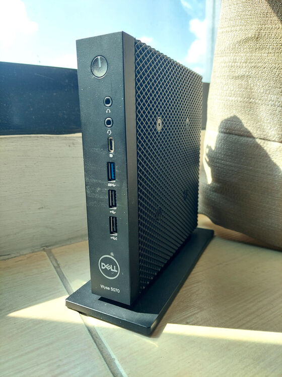 A Dell Wyse 5070 thin client computer next to a window. It is a small, slim, black PC, a vertical box about 7 inches tall and 7 inches deep, and only about 1.5 inches thick, mounted on a plastic base. You can see the power button and various ports on the narrow front panel, along with the Dell logo. The side panels have a grid-pattern surface structure that makes it look like a proper server computer, and probably helps with cooling the fanless system.